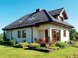 Cementowa dachówka w grafitowym kolorze na łamanym dachu kontrastuje z beżowym akrylowym tynkiem. Na poddaszu od strony ogrodu właściciele zdecydowali się na lukarnę, a nie na okno połaciowe. Pokój jest przez to bardziej ustawny i przestronny