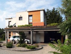 Wzdłuż frontowej i zachodniej elewacji rozciąga się wiata garażowa. Tylko jedno stanowisko parkingowe jest całkowicie osłonięte. Na dachu wiaty założono ogród z trawami i zbudowano drewniany taras. Właściciele lubią stamtąd patrzeć na zachód słońca