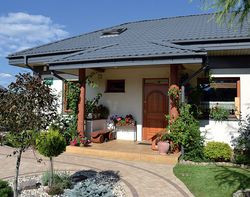 Ganek wykończono płytami z betonu imitującego piaskowiec. Bardzo dobrze się sprawdzają, ponieważ nie są śliskie. Daszek podpierają drewniane słupy, a ich rustykalny charakter podkreślają ławeczki. Podjazd i ścieżkę do domu utwardzono betonową kostką, ale dodano również zieleń, by przed domem było przytulniej