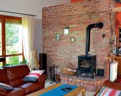 Ściana przy kozie grzewczej jest wykonana z cegły. Nawiązuje do niej kolor ścian w kuchni. Nad salonem, zamiast pierwotnego stropu, zastosowano dwa solidne podciągi, a przestrzeń do stropodachu pozostawiono otwartą. Połączone pomieszczenia dzienne są dzięki temu wysokie i przestronne