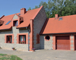 Rozłożysty dom umieszczono w głębi posesji pod ścianą lasu. W skrzydle północnym znajduje się – garaż, pośrodku – strefa dzienna i rodzinna. Elewacje wykonano z szarej okładziny betonowej, imitującej łupany kamień oraz z czerwonej pełnej cegły klinkierowej. Dach pokryto ceramiczną dachówką