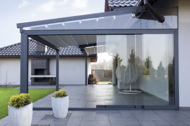 Pergole z proszkowo malowanego aluminium, na których zamocowano rozkładany dach nad tarasem z odpornego na wodę, trudno zapalnego włókna