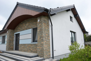 Zimną kolorystykę elewacji przełamują ciepłe tony cieniowanego kamienia elewacyjnego oraz podbitki dachowej z PVC