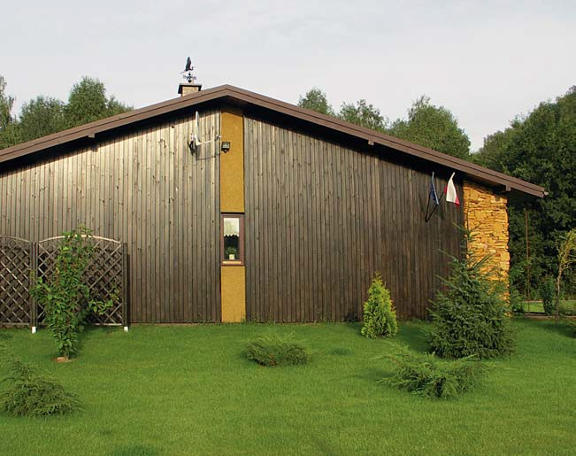 Na bocznych ścianach budynku, wyłożonych okładziną z drewna, celowo pozostawiono pionowe pasy tynku, biegnące od szczytu aż po podmurówkę. Tego typu wstawka rozbija jednolitą ciemnobrązową ścianę i optycznie ją wyłduża. Podobny efekt dają ustawione pionowo deski oszalowania