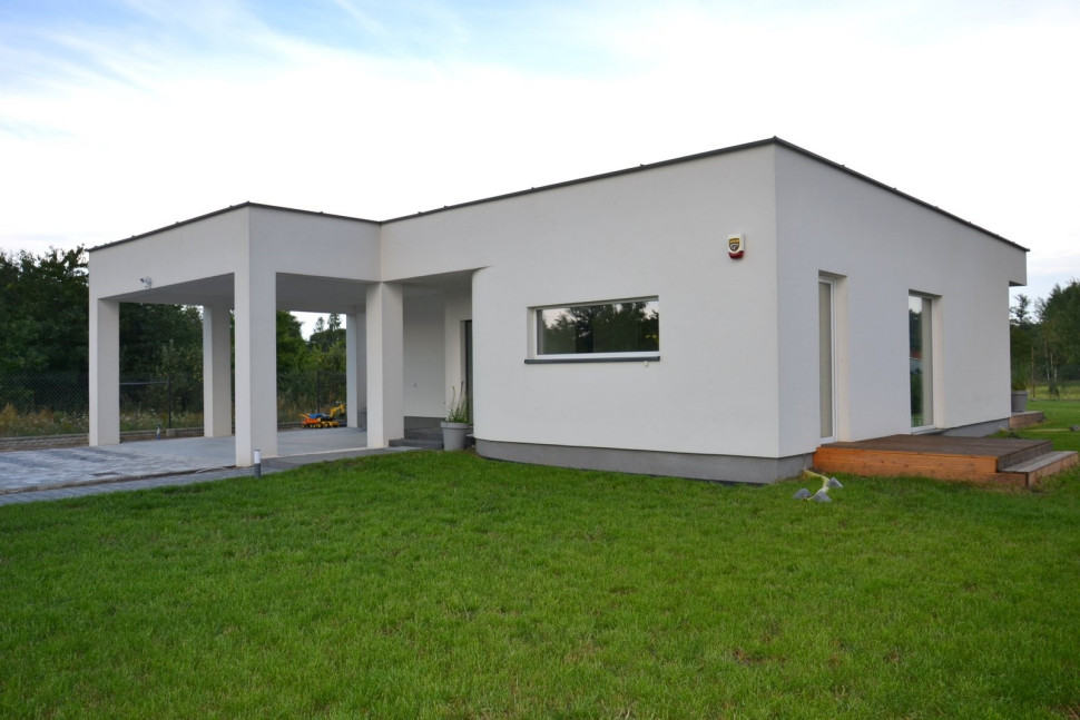 Parterowy budynek z płaskim dachem, o minimalistycznej bryle w kolorach bieli i szarości z dodatkiem naturalnego drewna