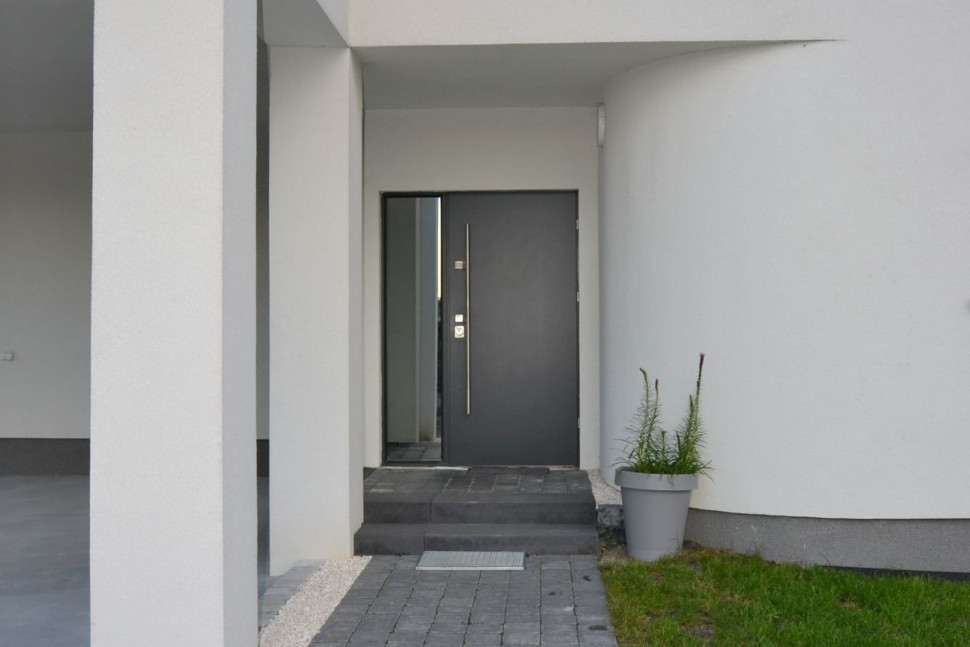 Aluminiowe drzwi wejściowe ulokowano we wnęce, chroniącej całe wejście przed opadami atmosferycznymi i wiatrem