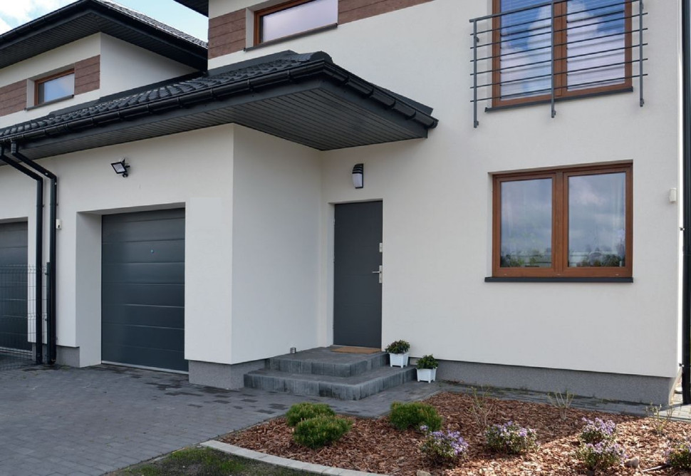 Główne wejście znajduje się pod wysuniętym okapem dachu, przykrywającym garaż na jeden samochód