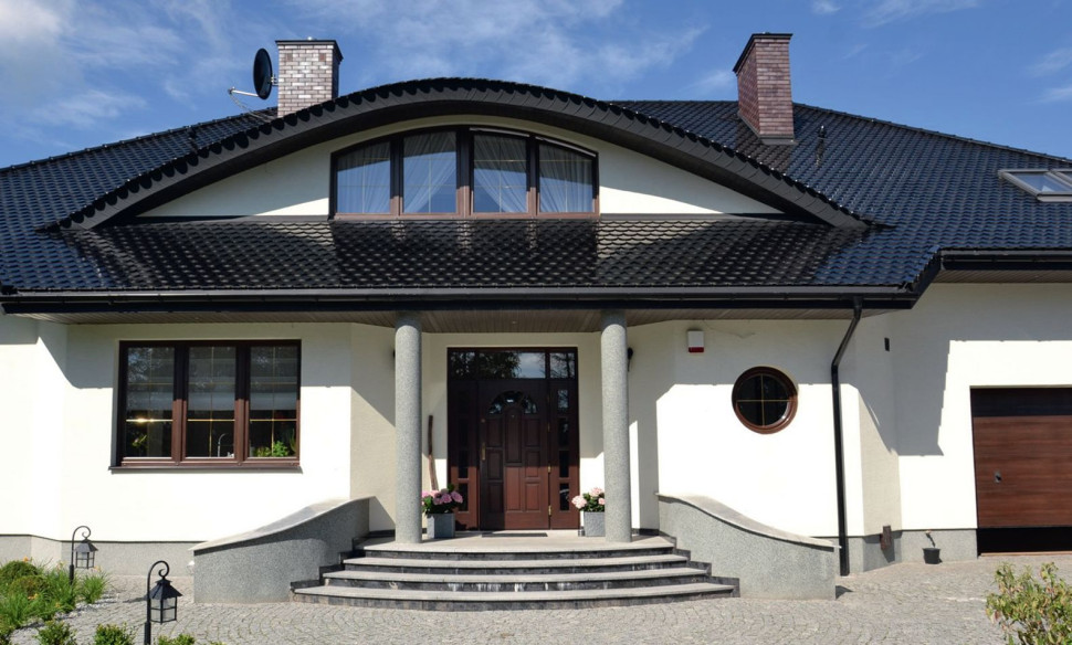 Główne wejście do budynku jest szerokie i zamknięte efektownymi drewnianymi drzwiami z górnym i dwoma bocznymi doświetlami