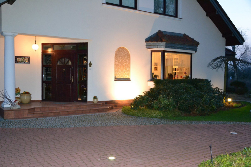 Oświetlenie na froncie domu tworzą wisząca lampa na ganku, halogen podświetlający fresk na elewacji, niska latarenka przy narożniku domu oraz oprawy najazdowe w bruku na rondzie