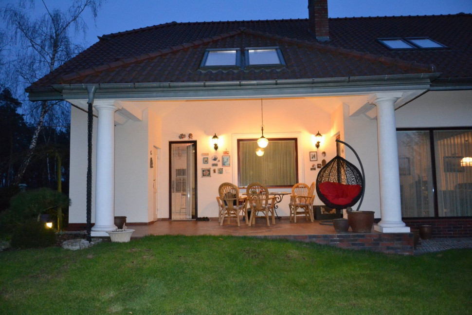 Kinkiety i wisząca lampa ciepłym światłem oświetlają zadaszony taras