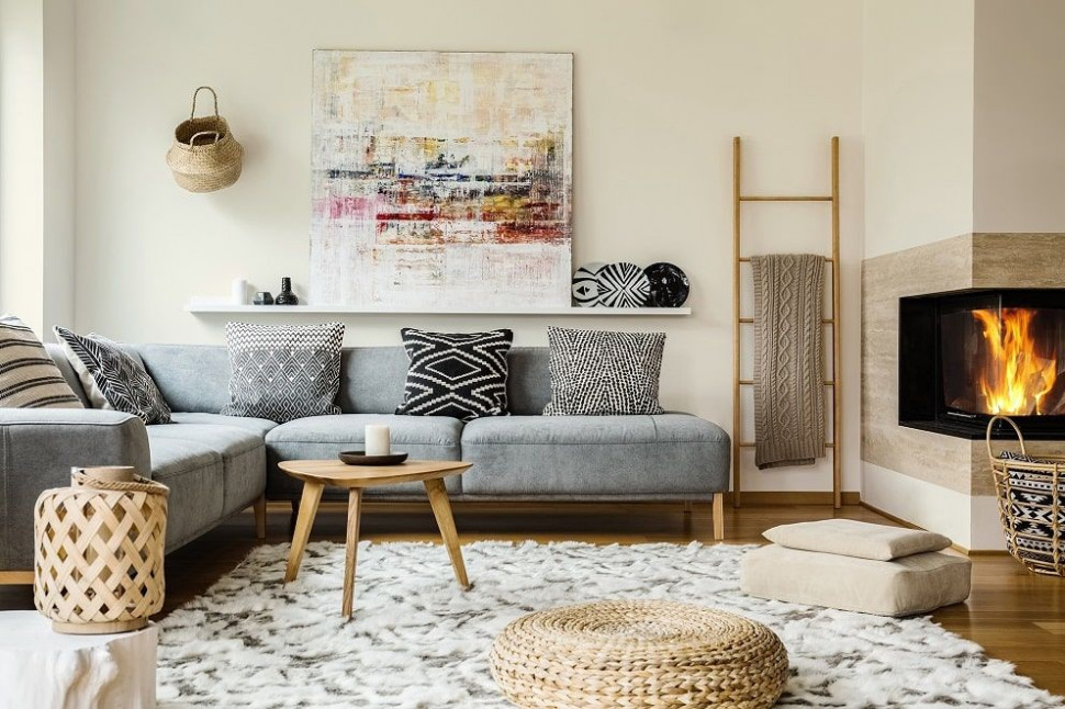 Rustykalny salon w bloku: Szara sofa, drewniany stolik kawowy, pleciony puf, drabinka jako wieszak, dekoracje ocieplające aranżację. Kominek w tradycyjnym stylu, nowoczesny obraz na ścianie, farba Śnieżka Barwy Natury w kolorze 101 Posrebrzane Żyto.