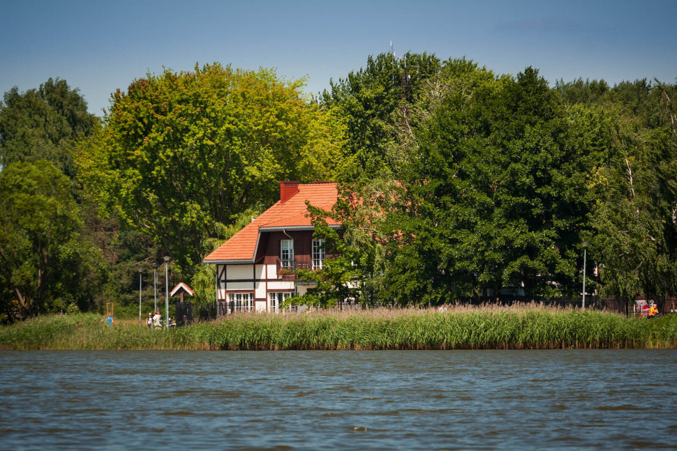 Dom nad jeziorem z ogrodzeniem i trzcinami przy brzegu, otoczony drzewami, widziany od strony wody