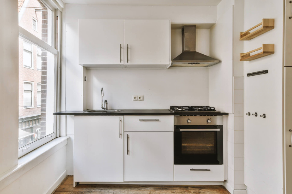 Mała biała kuchnia w bloku z oknem, zlewem, piekarnikiem i półkami na ścianie